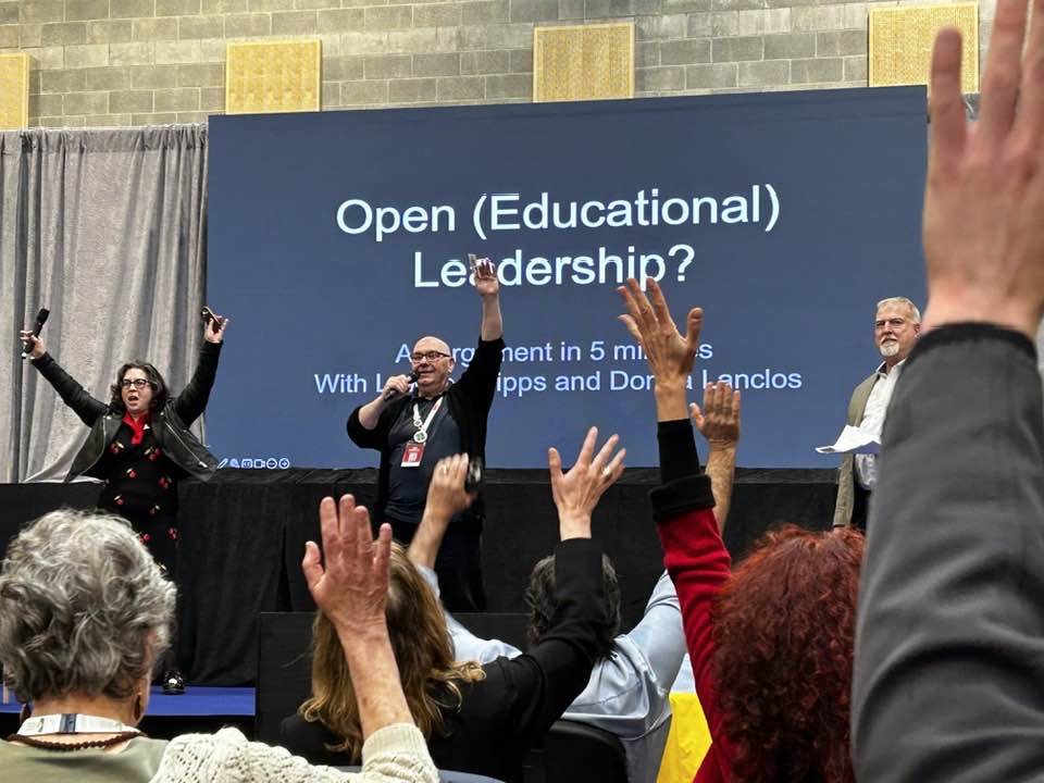 Presenters on a Stage waving hands in front of a crowd. A Slide on a screen reads "Open (Educational) Leadership?" 